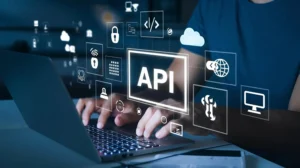 APIs: O Que são e como elas fazem diferentes sistemas conversarem: Pessoa usando notebook com interfaces e ícones digitais ao redor, com destaque para o termo “API” em um painel, simbolizando comunicação entre sistemas e integração via APIs. Ao fundo, nuvem e segurança em elementos gráficos