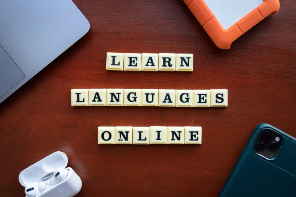 Cursos de Idiomas: Domine Novas Línguas: Cursinhos de Idiomas: Domine Novas Línguas ilustrado com palavras em blocos que formam “LEARN LANGUAGES ONLINE”, além de notebook, celular e fone de ouvido sobre uma mesa de madeira.