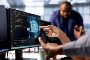 Equipe de tecnologia em escritório trabalhando com código em um monitor, enquanto uma pessoa aponta para o código, mostrando desenvolvimento de software e colaboração em projetos de TI. Low-Code e No-Code: Como essas tecnologias estão facilitando o desenvolvimento