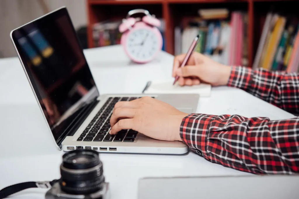 Mãos digitando em um notebook em uma mesa de estudo com livros ao fundo e relógio na mesa, enquanto uma pessoa trabalha ou estuda; Como Escolher Um Computador Adequado Para Trabalho, Estudos Ou Uso Profissional